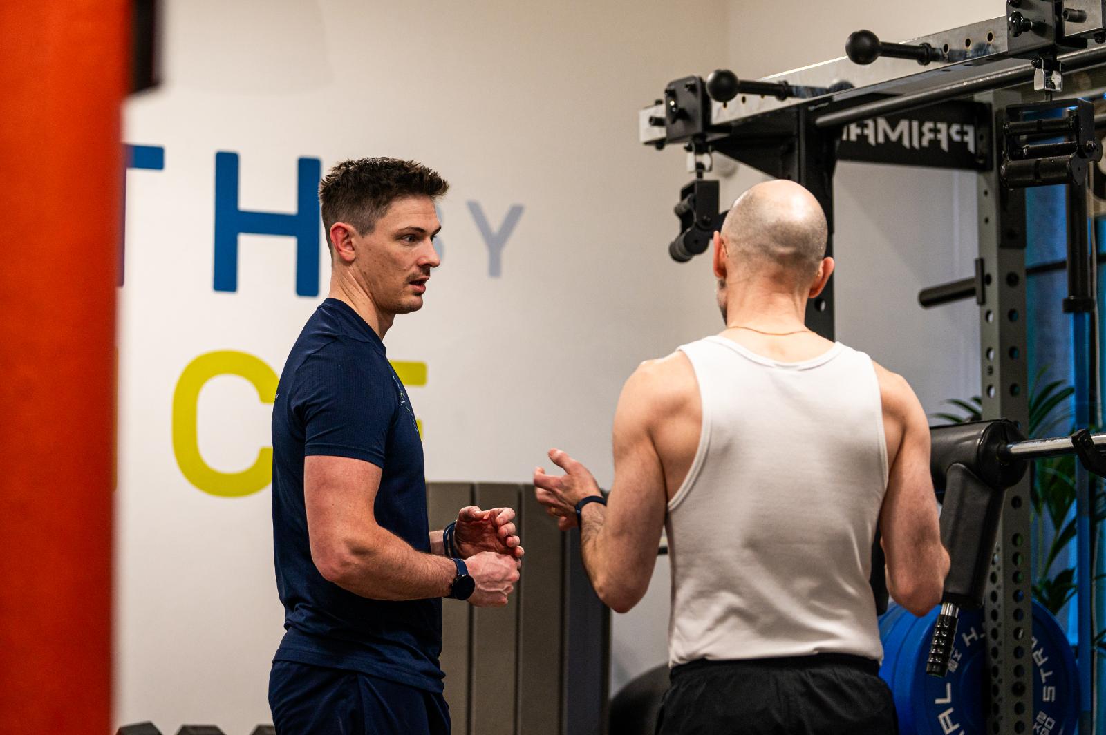 A Health By Science coach in a dark blue uniform discussing a seamless transition from rehab to strength training with a male client in a white tank top.