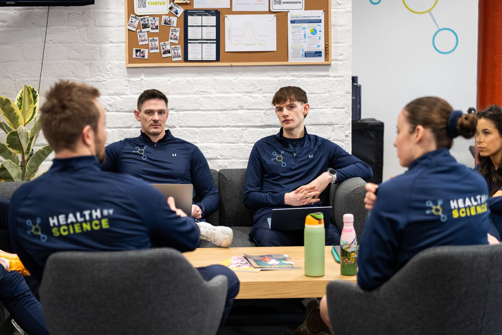 Four members of the Health By Science team wearing matching blue branded fleeces, sitting together in a bright office area having a collaborative discussion.