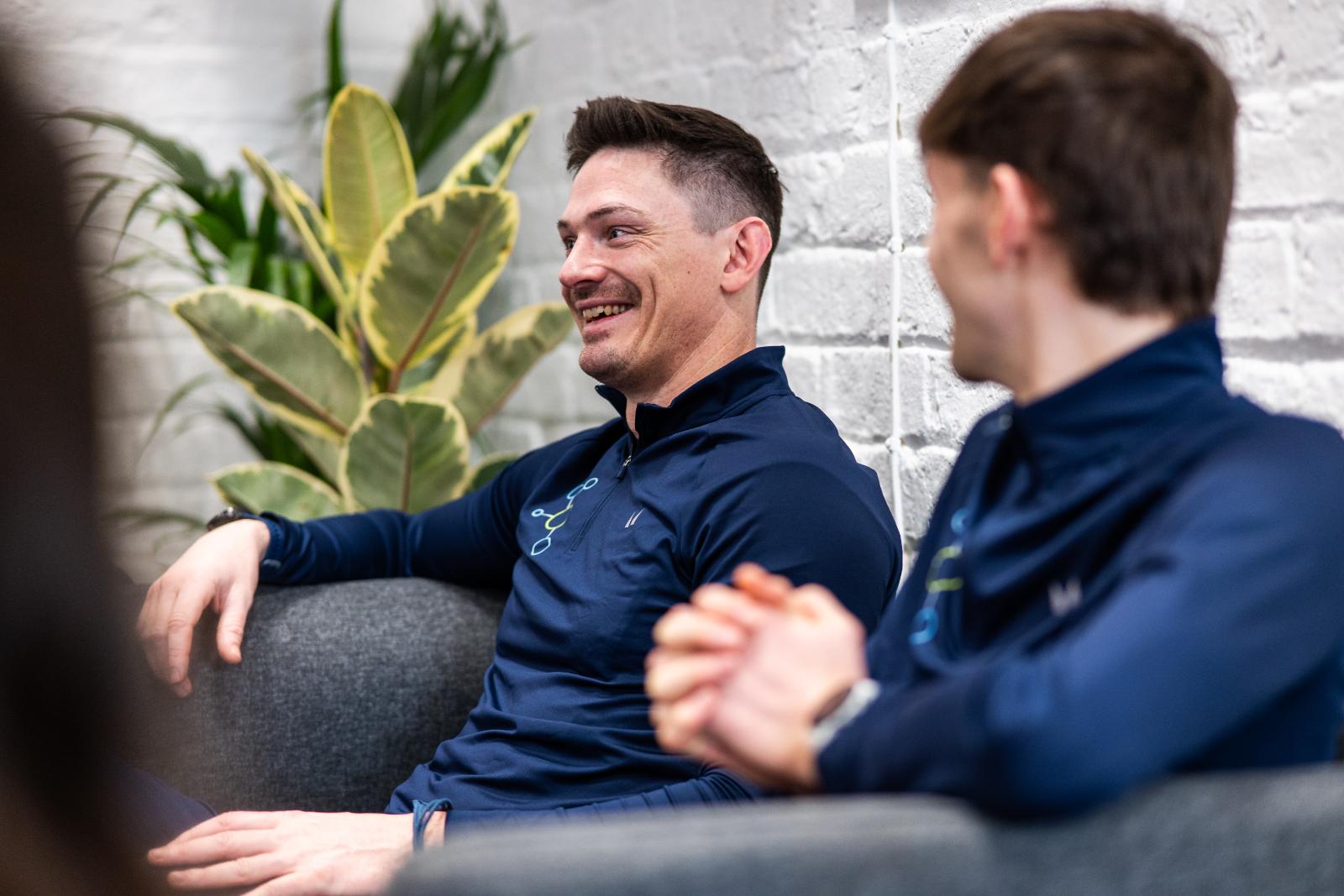 Two male members of the coaching team smiling and talking while sitting in the clinic lounge area.