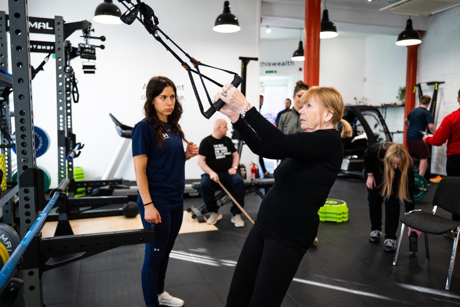A woman performing a guided suspension trainer (TRX) exercise under the supervision of a rehab coach.