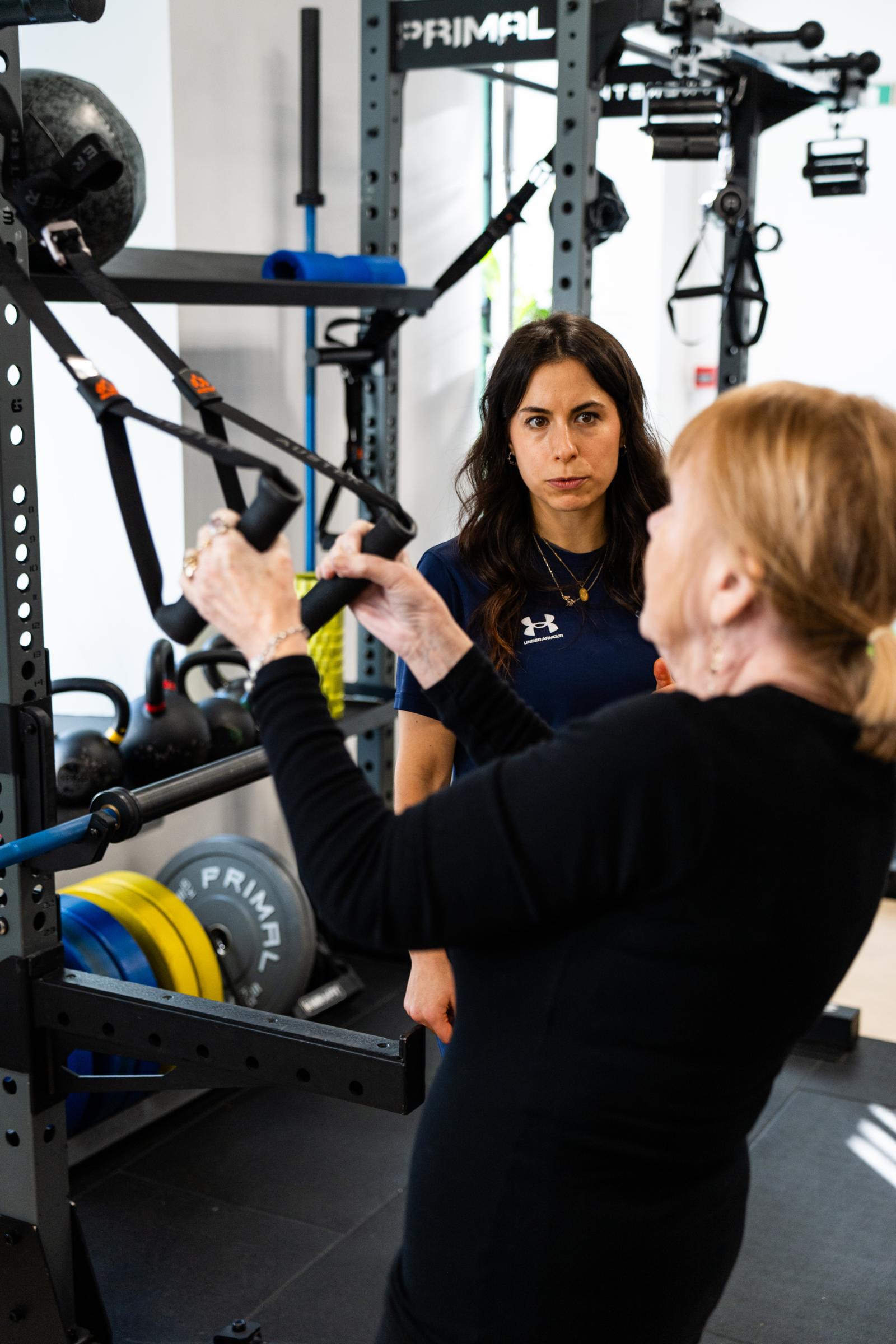 Health by Science coach monitoring movement mechanics during a clinical rehabilitation session using suspension training equipment.