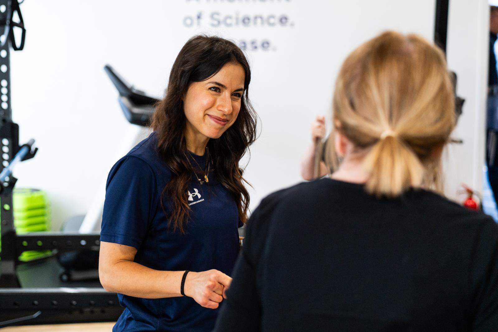 A smiling female rehab coach talking to a client in a bright, modern fitness studio.