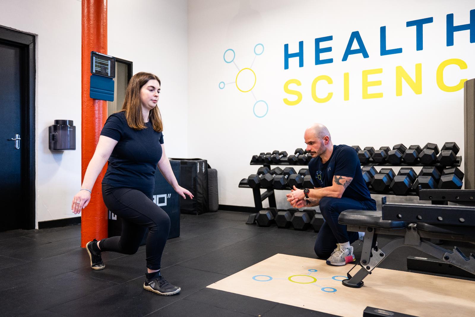 Female athlete performing a reverse lunge with a personal trainer in a gym setting.