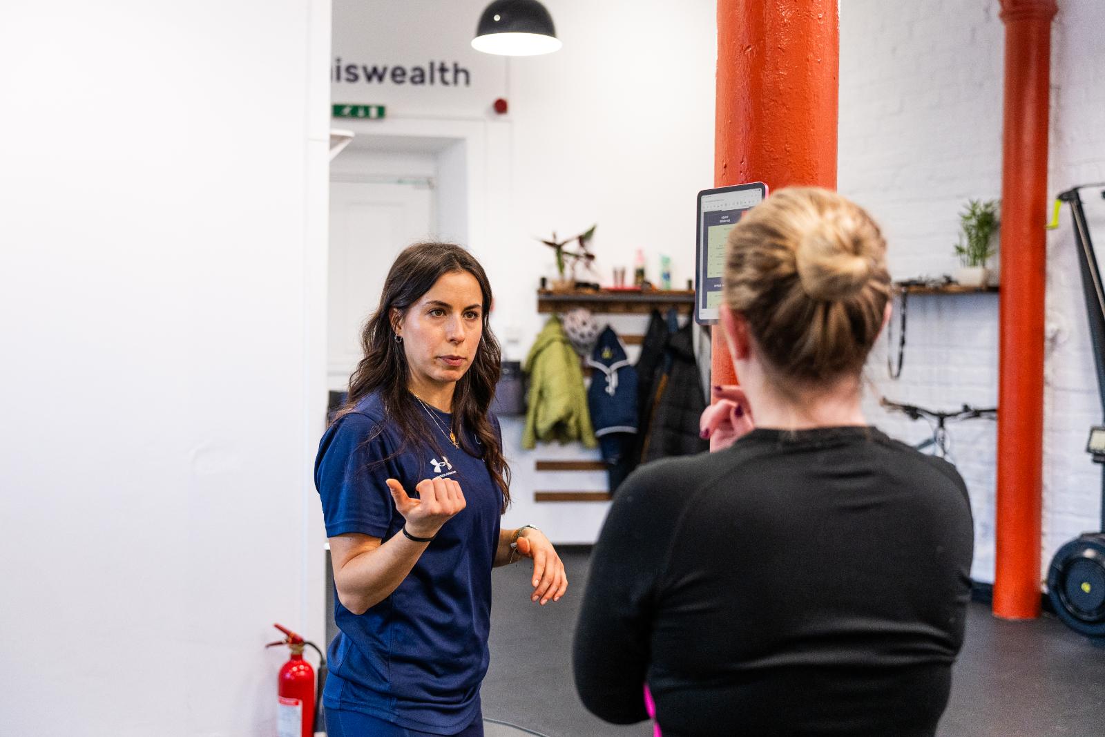 A female clinician wearing a blue polo shirt explaining a movement plan to a client in the gym.