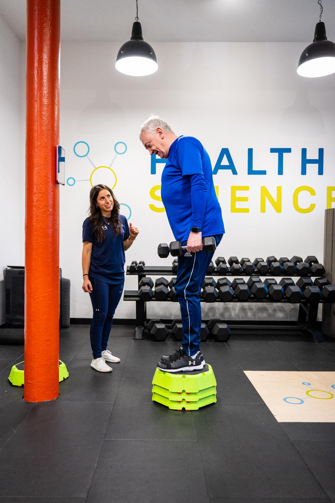 A female Health By Science physiotherapist talking with an older male client who is holding a dumbbell, demonstrating a supportive and inclusive training environment.