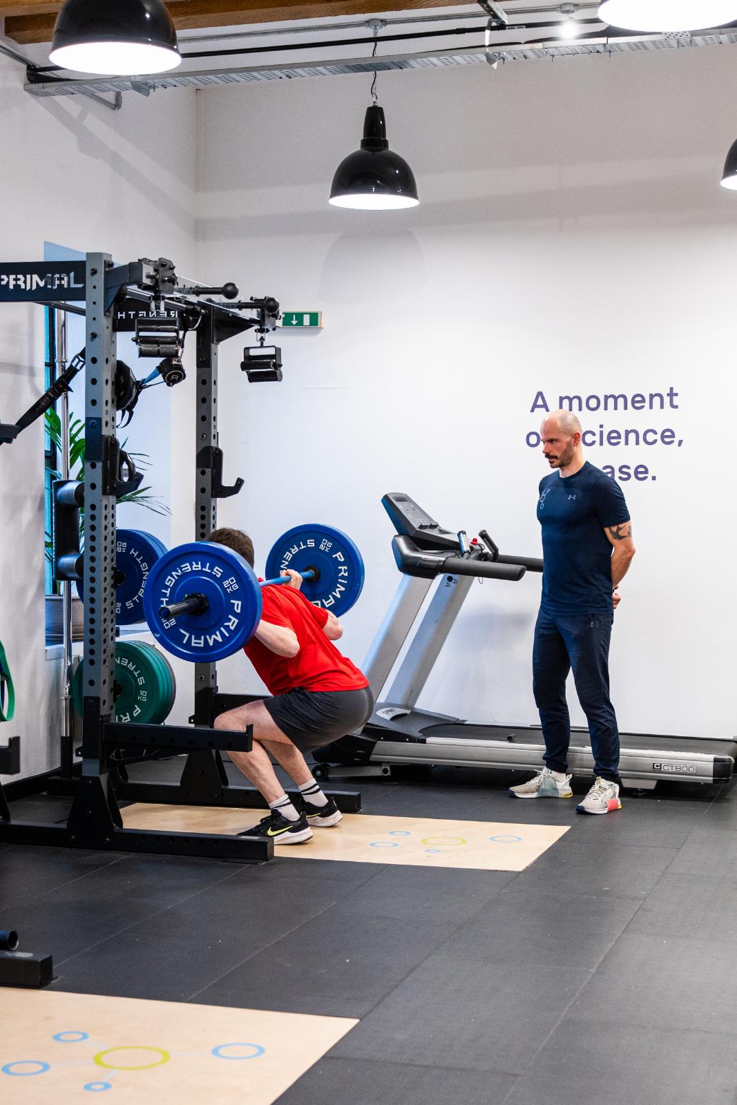 Clinical specialist monitoring barbell squat depth and spinal alignment for a personal training Edinburgh client.