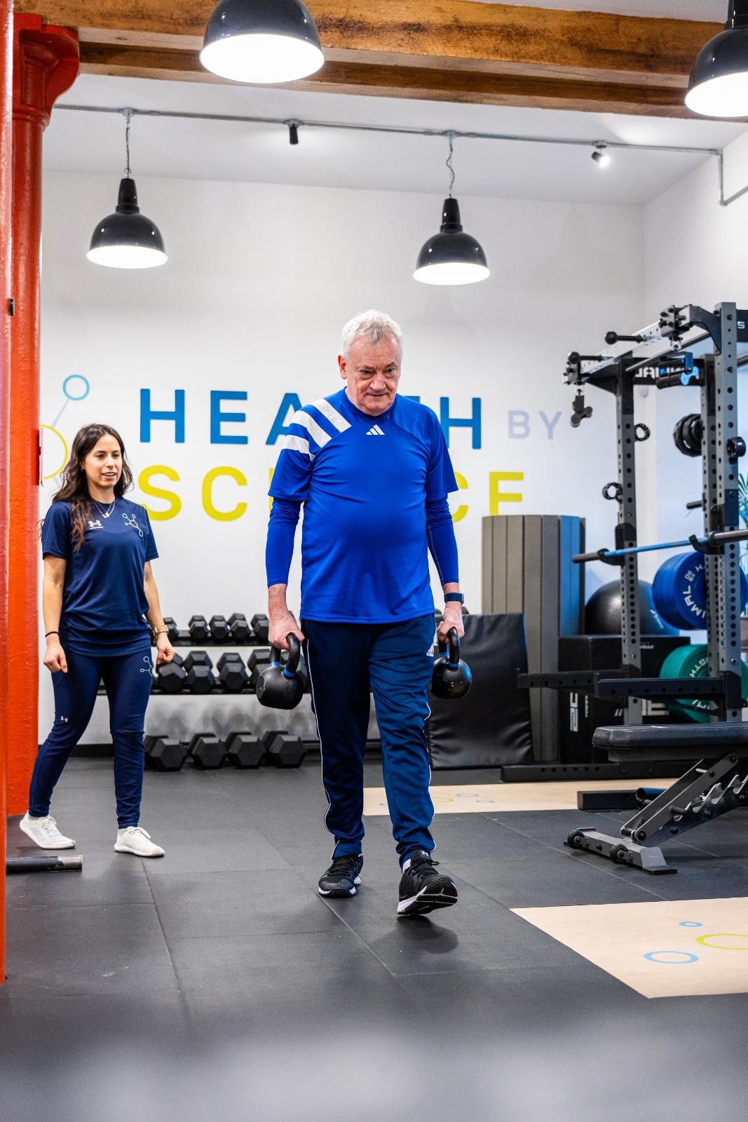 Health by Science coach supervising a patient performing a farmers carry with kettlebells to build functional grip and core strength.