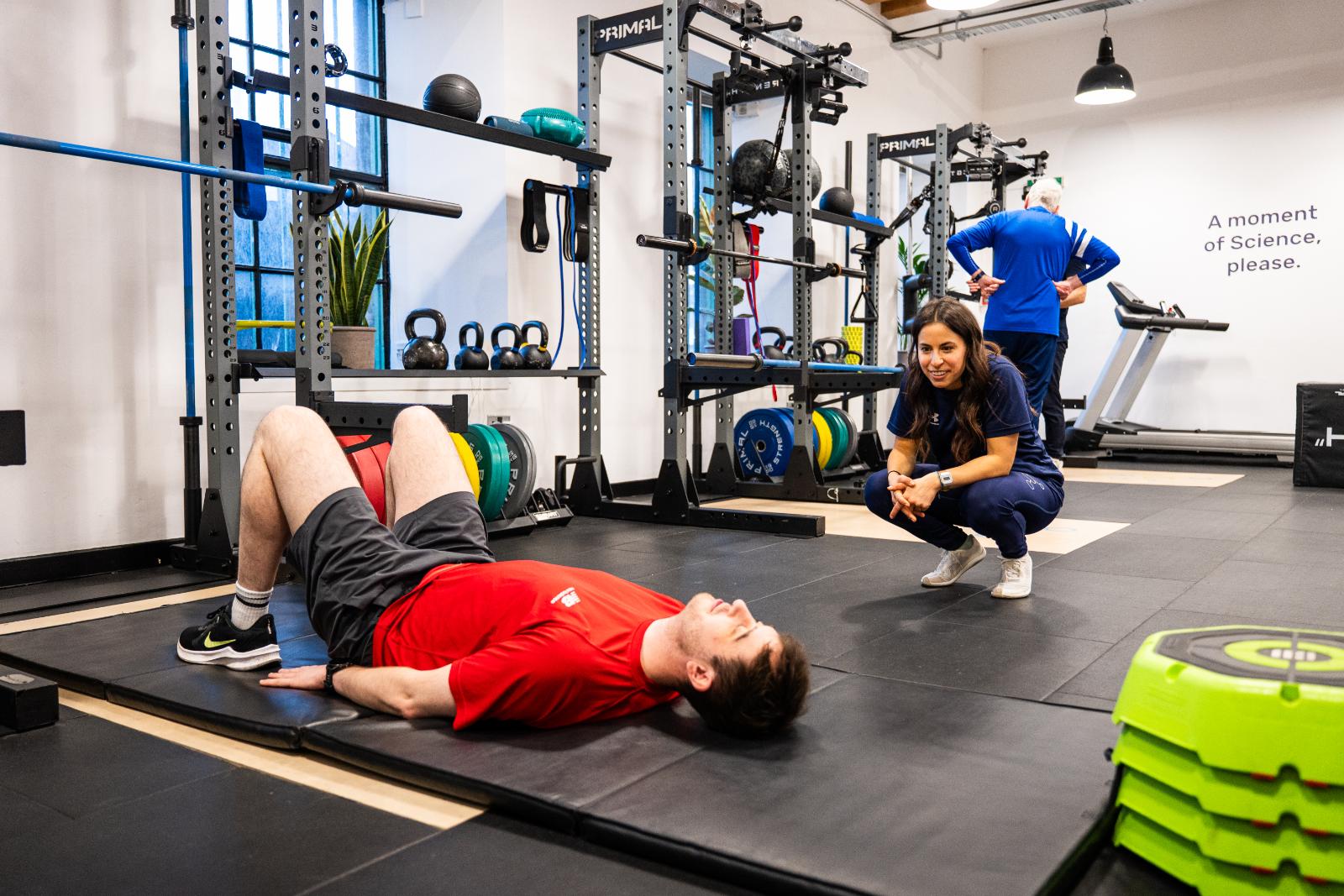 A male client lying on a gym mat performing a bridge exercise while a female trainer crouches nearby to assess his movement and form.