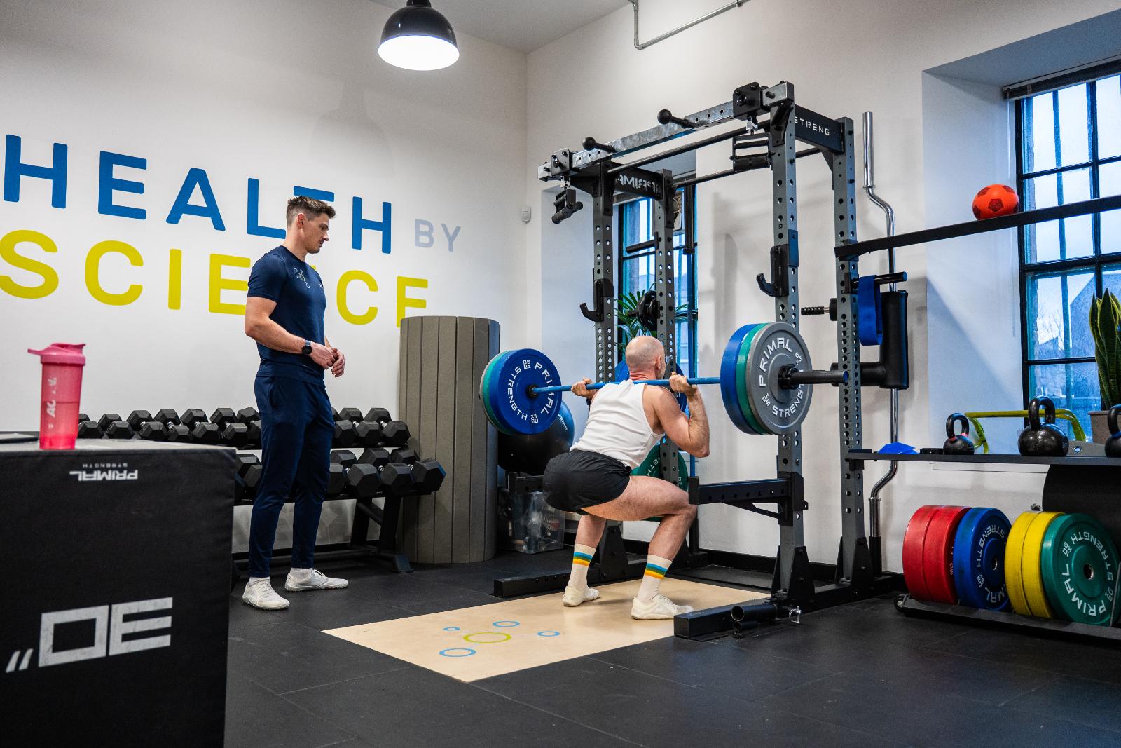 A man performing a heavy barbell back squat in a high-performance gym while a coach monitors his form.