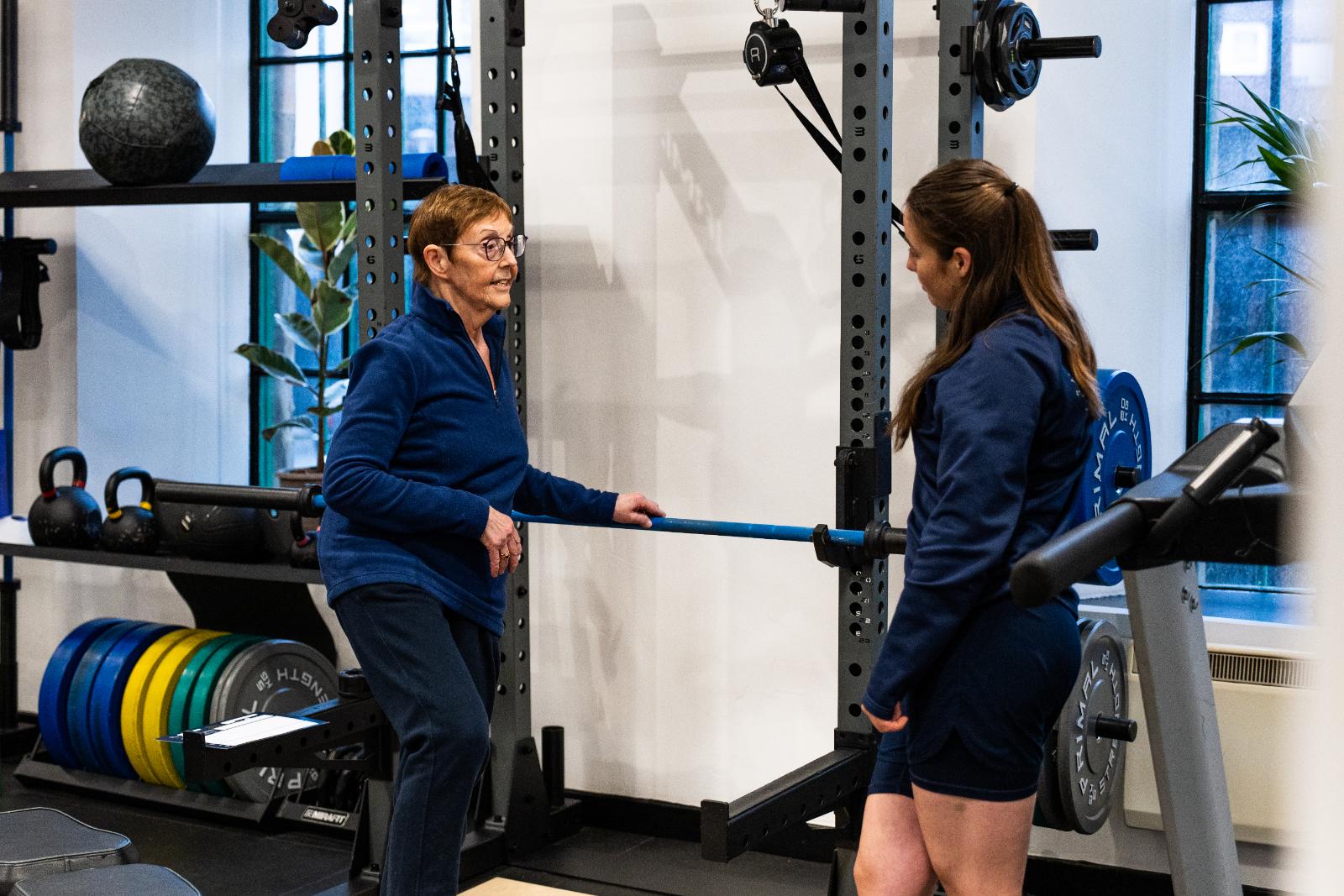 Professional consultation between a Health by Science coach and a senior patient regarding safe barbell exercise progressions.