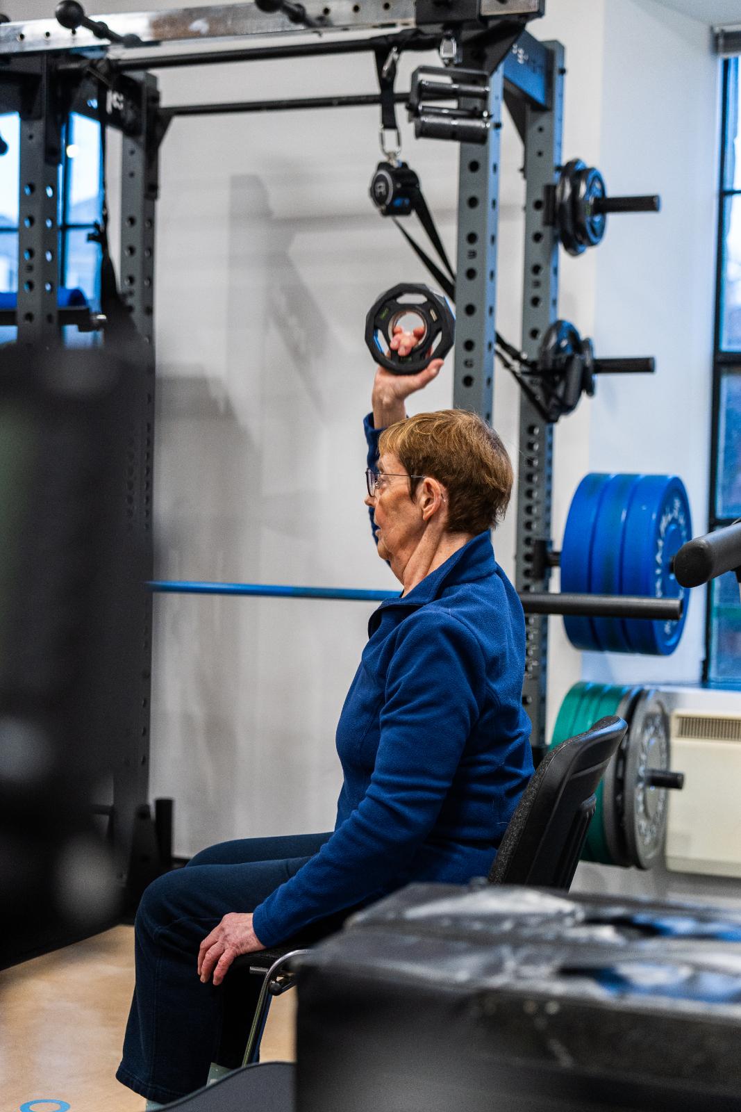 Specialist monitoring a seated single-arm overhead press for an older adult to assess upper body strength and joint health.