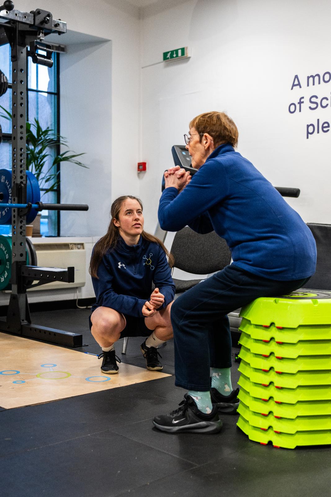 Health by Science coach monitoring a box squat progression to assess knee tracking and hip mobility for a senior patient in Leith.