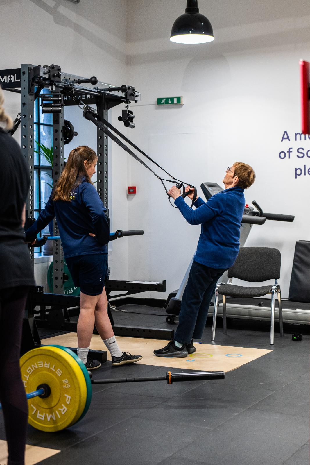 Specialist coach monitoring a suspension trainer row to assess upper body pulling mechanics for a senior rehabilitation client.