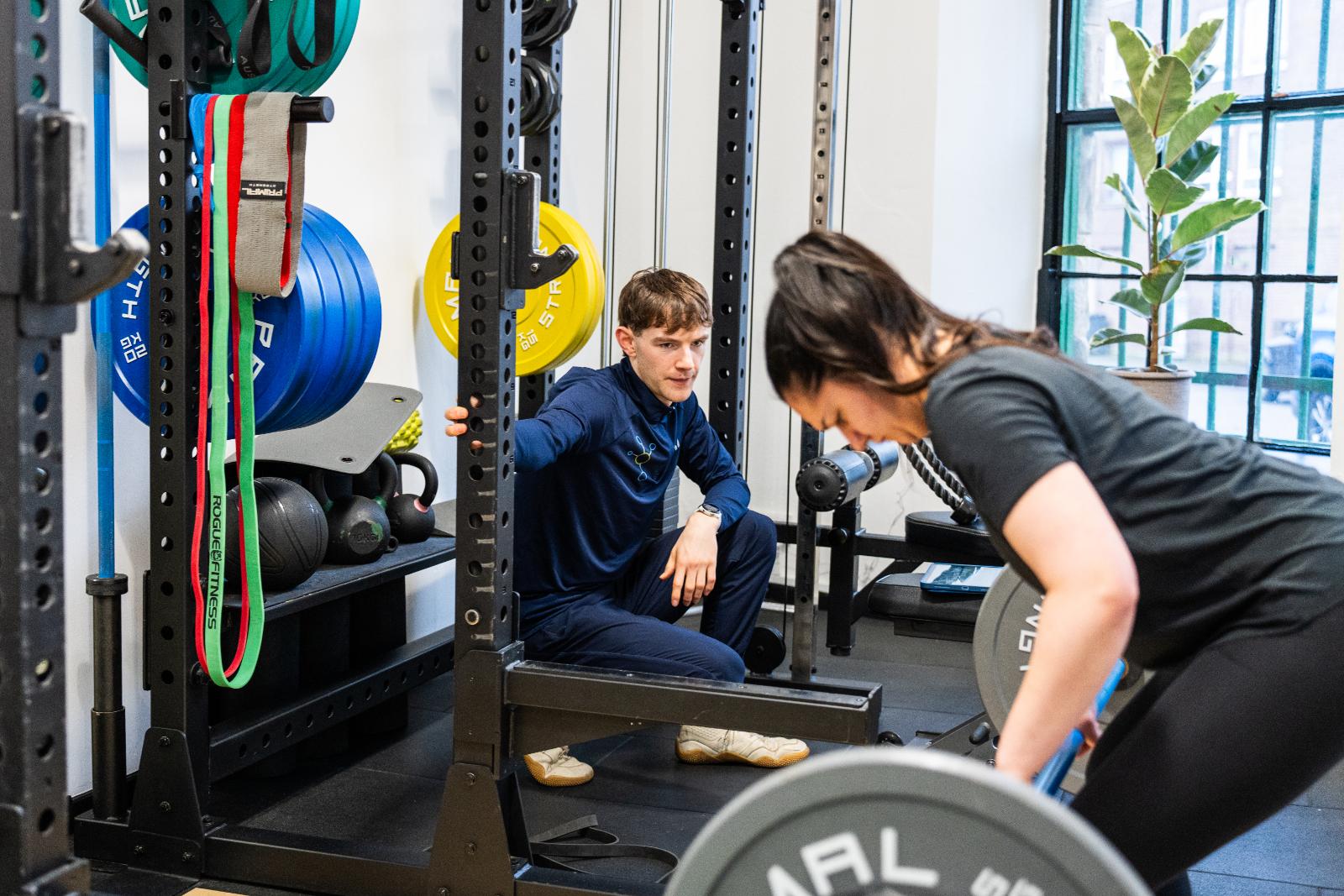 A male personal trainer monitoring a client's form during a barbell lift in a structured gym setting.