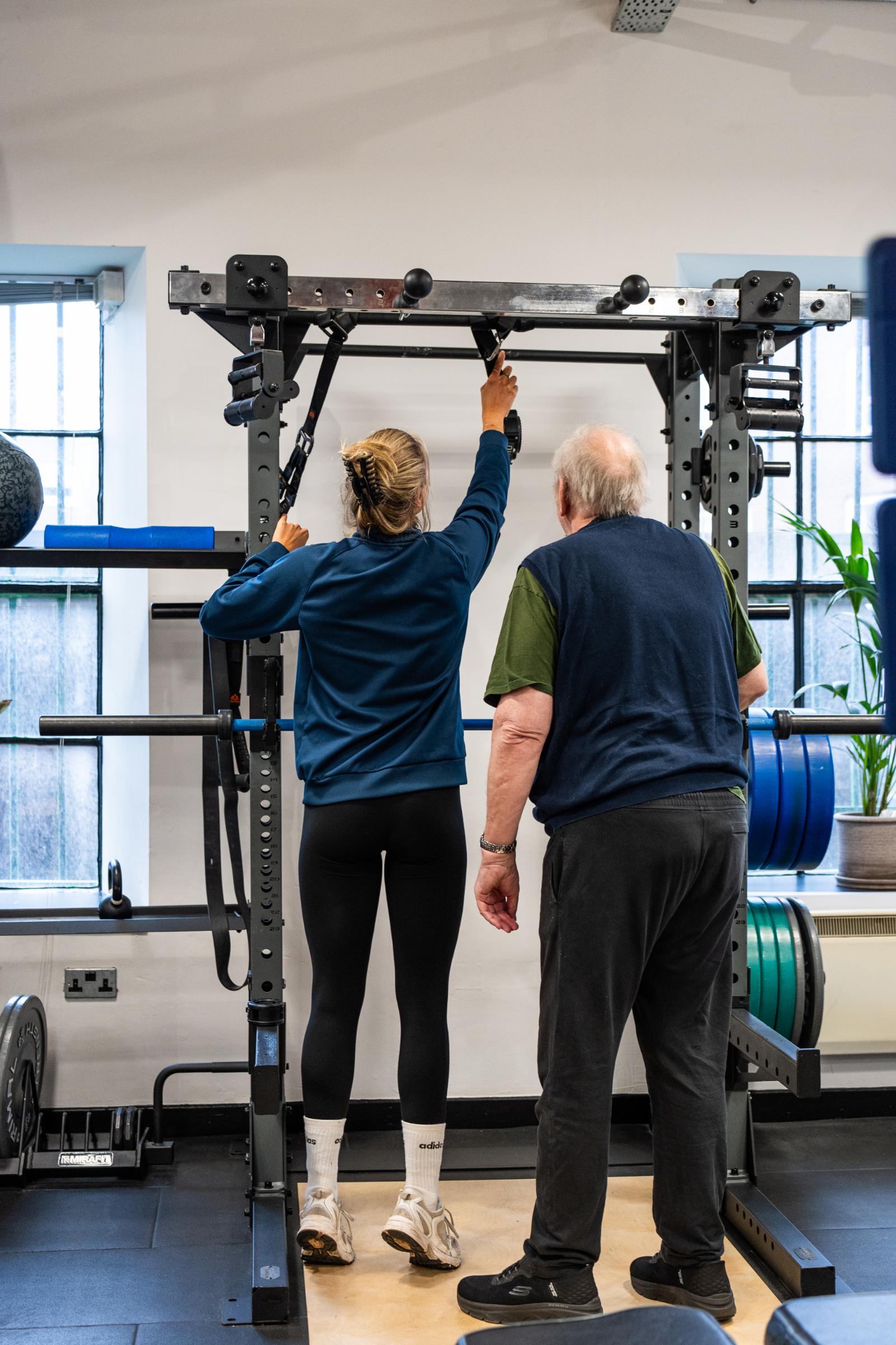 Health by Science specialist providing clinical assistance to a senior patient while adjusting height on a functional training rig.