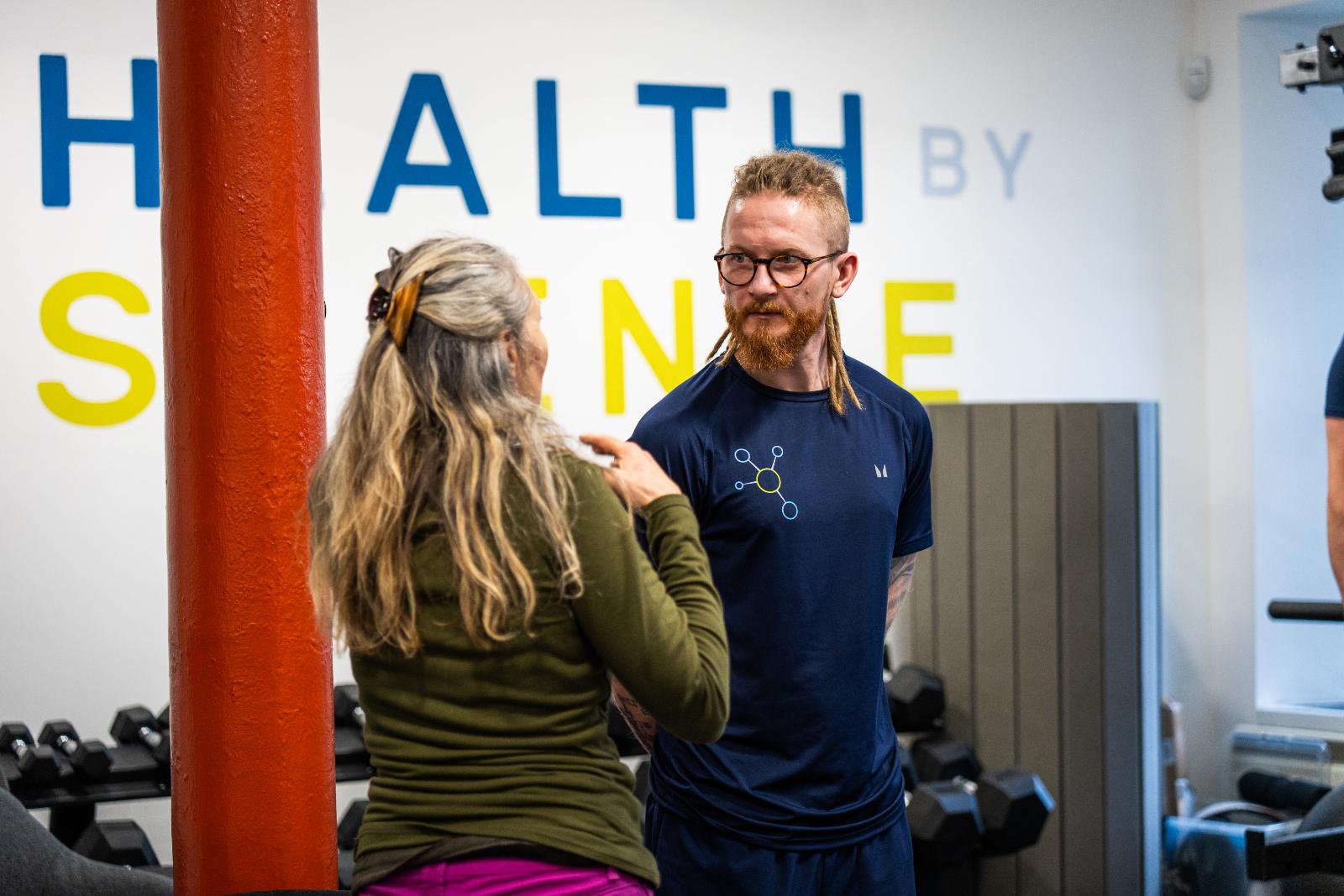 A rehab coach in a blue Health By Science uniform chatting supportively with a client during a supervised session.