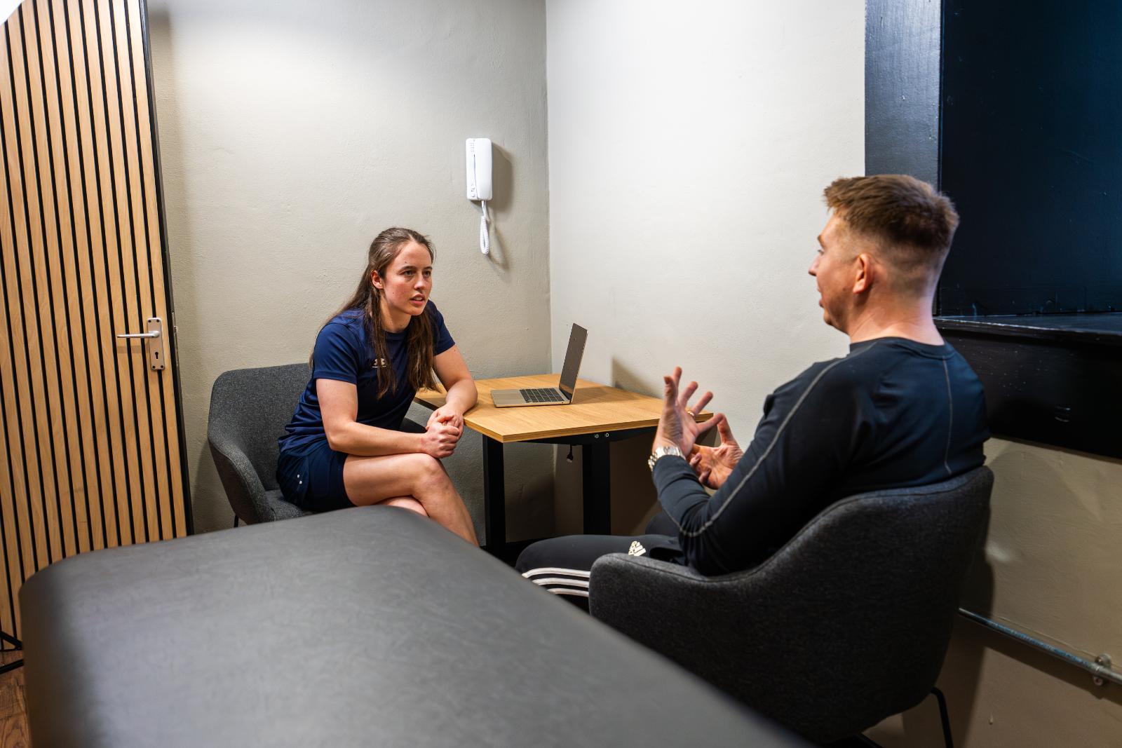 Two people seated in a physiotherapy clinic discussing a rehabilitation plan and injury diagnosis.