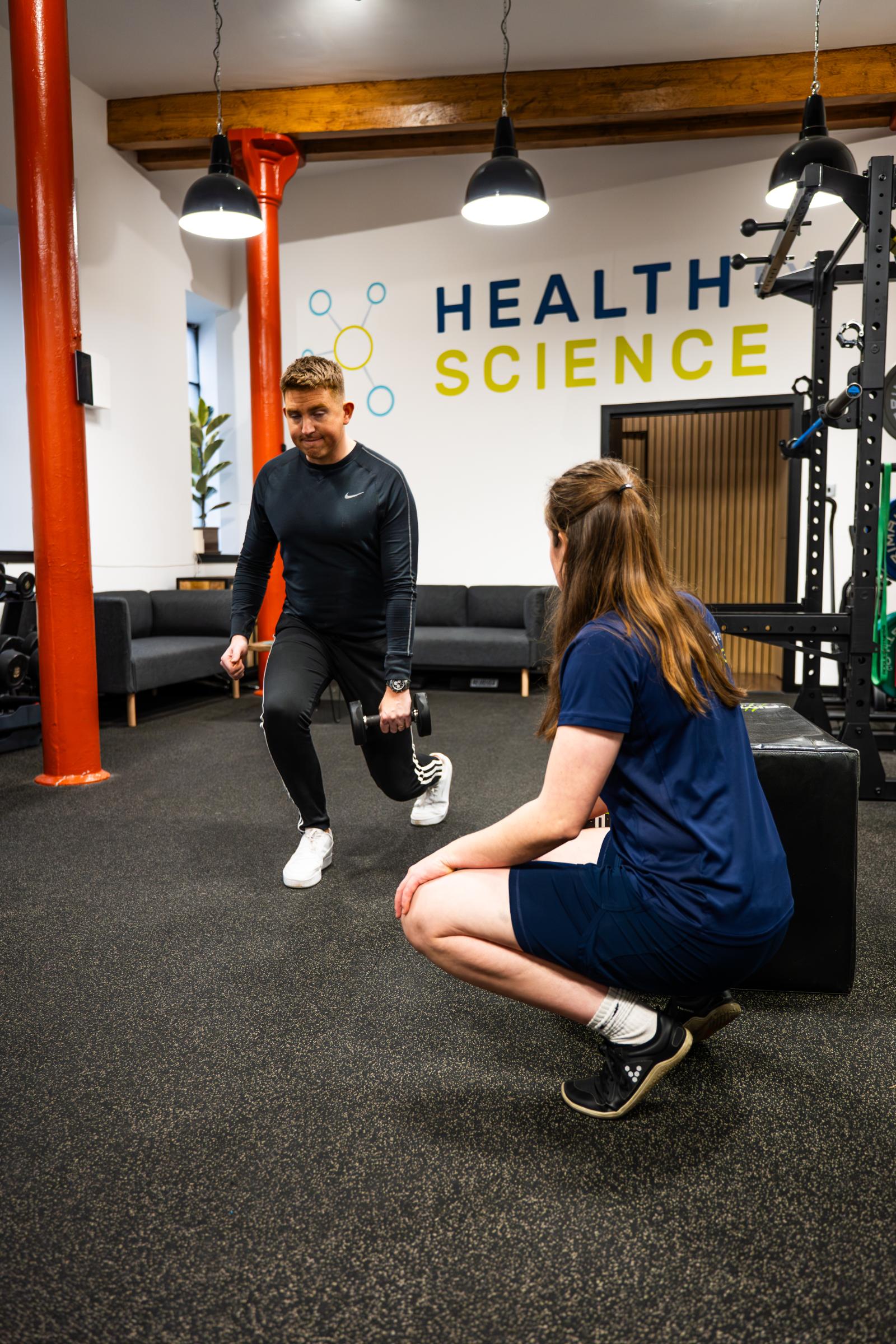 Health by Science specialist monitoring a weighted reverse lunge to assess lower body stability and unilateral strength for a rehabilitation patient.