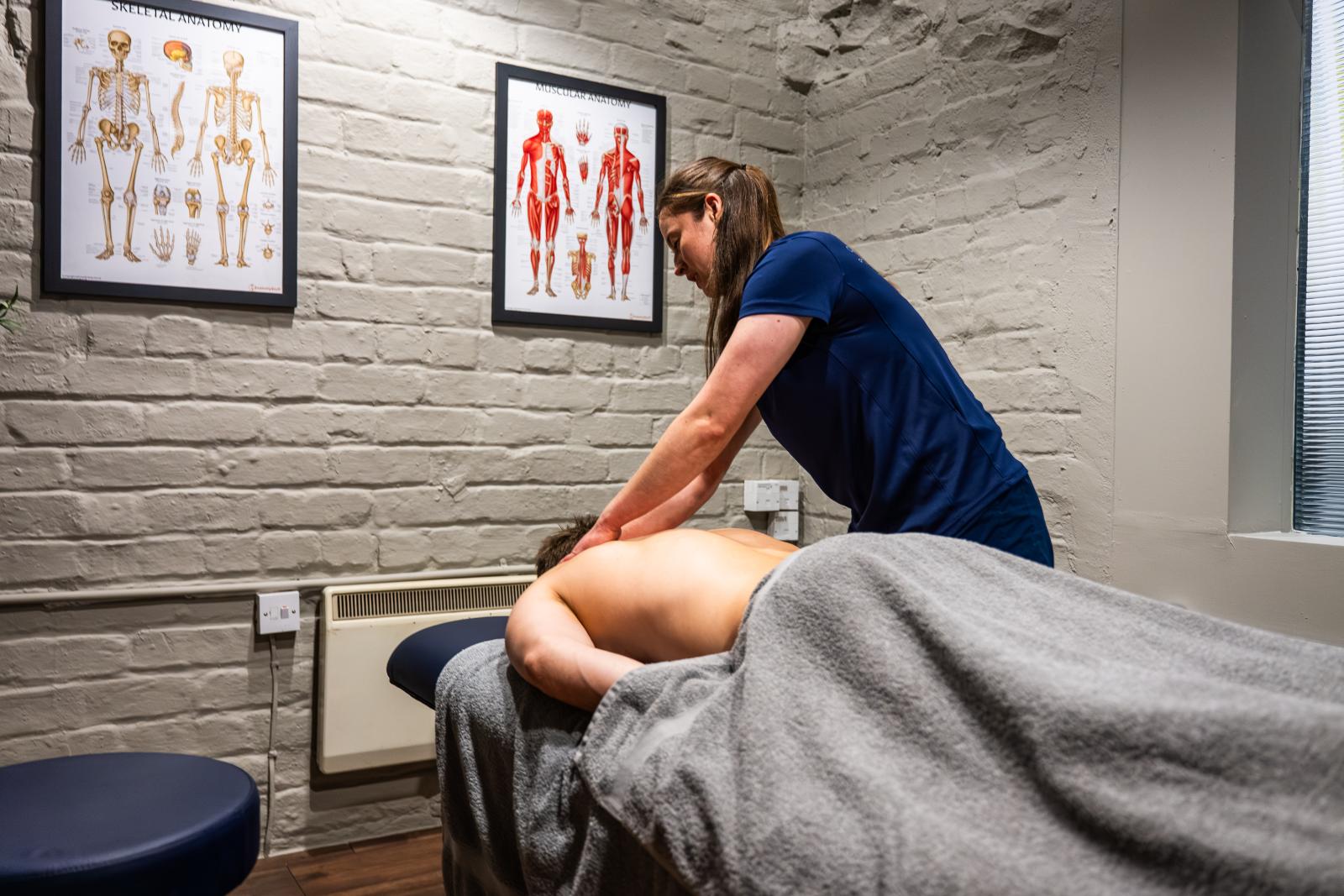 A female therapist performing a sports massage in a clean, professional clinic room with anatomical posters in the background.