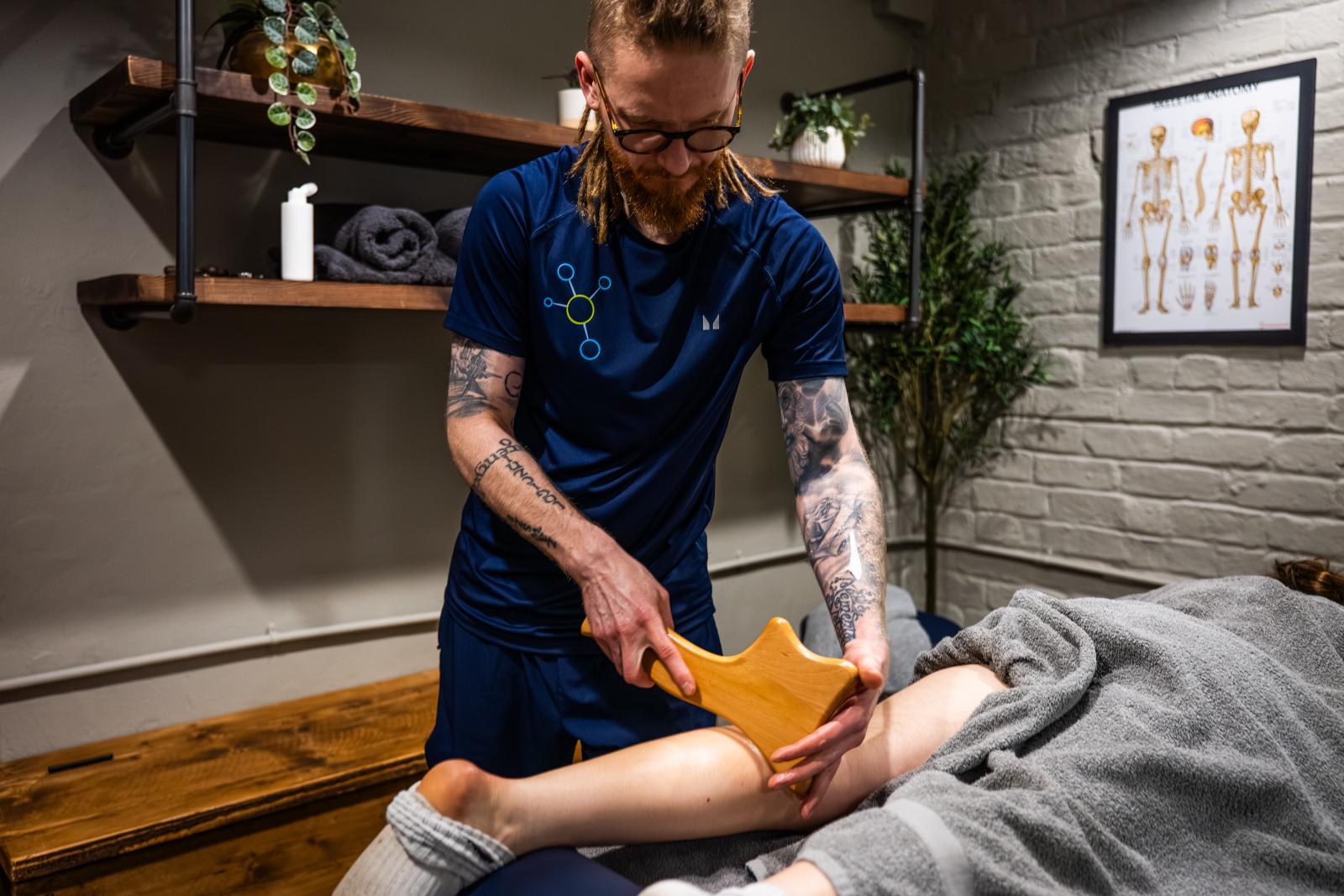 A male therapist using a specialized wooden massage tool for deep tissue muscle recovery on a patient's leg.
